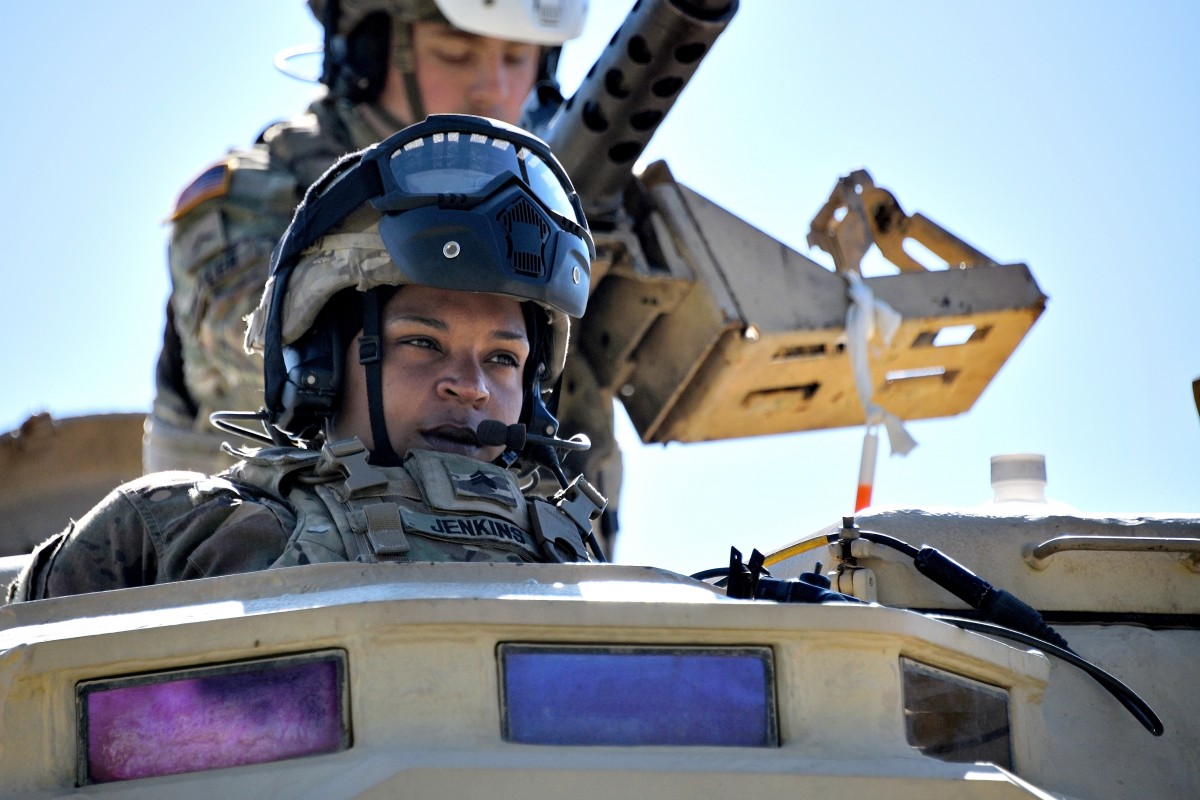 A servicemember peers out from inside a vehicle with another behind them at a mounted gun